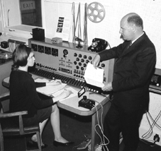 Delia Derbyshite seated with pen, Desmond Briscoe on the right staring down at her